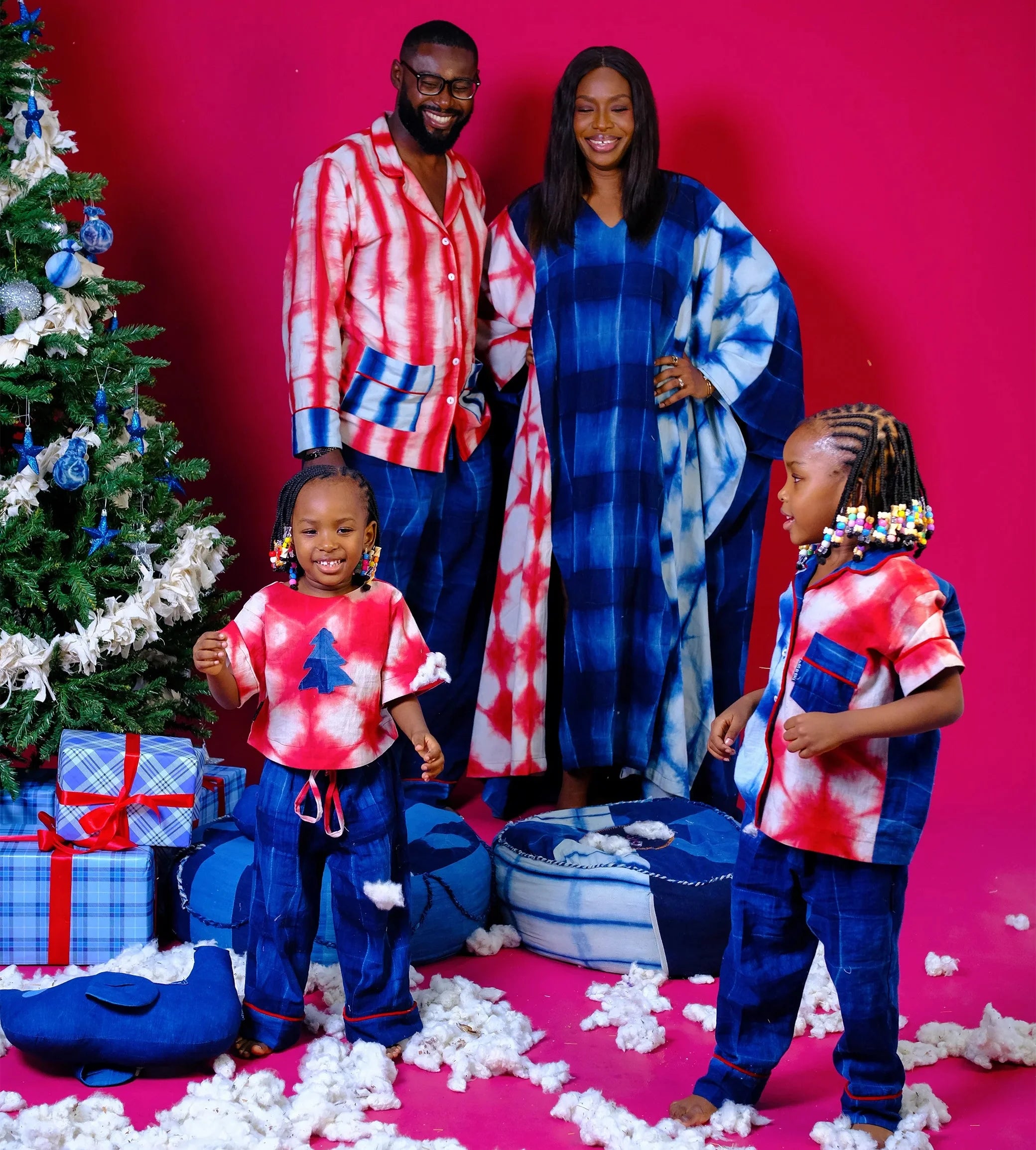 Family of four wearing tie-dye outfits in front of a Christmas tree and presents on a pink background